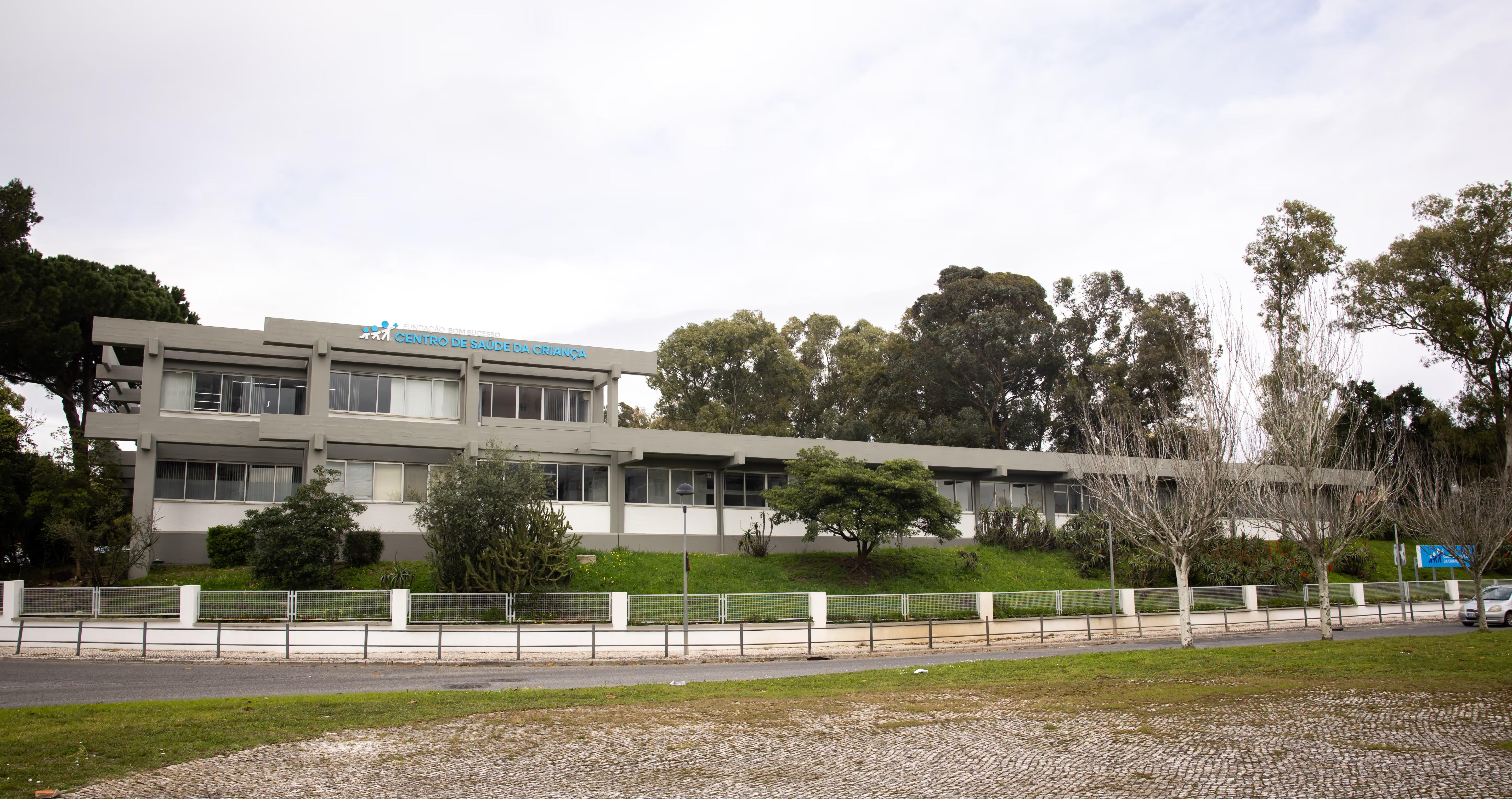 Modern two-story health center building with trees and a road in front under a cloudy sky.