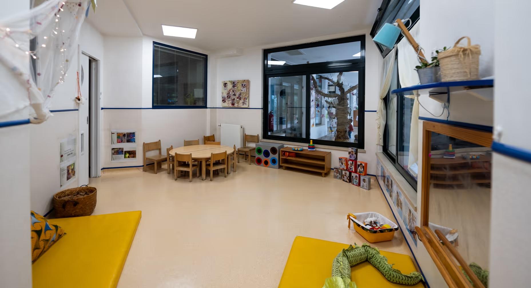 Bright preschool classroom with round table, wooden chairs, toys, mats, and a large window showing a tree decoration.