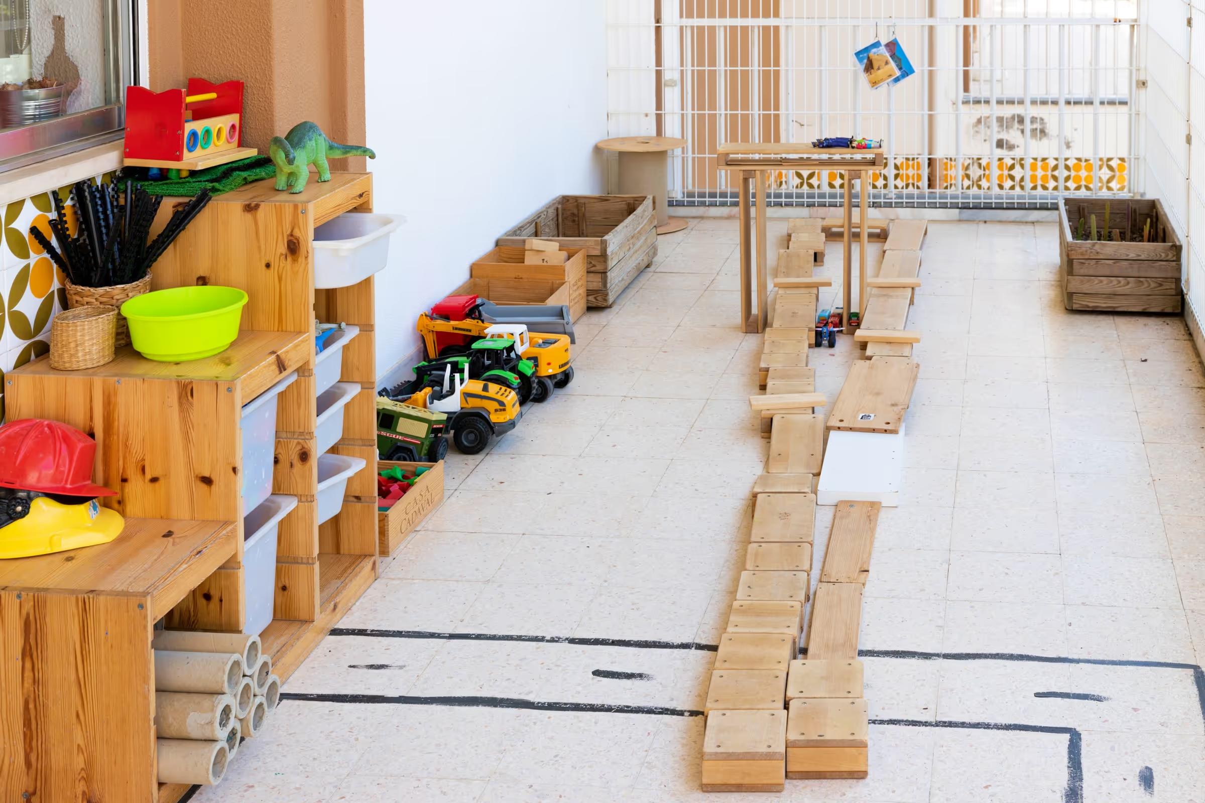 Play area with wooden shelves, toy trucks, dinosaur, helmets, and a wooden race track on the floor.