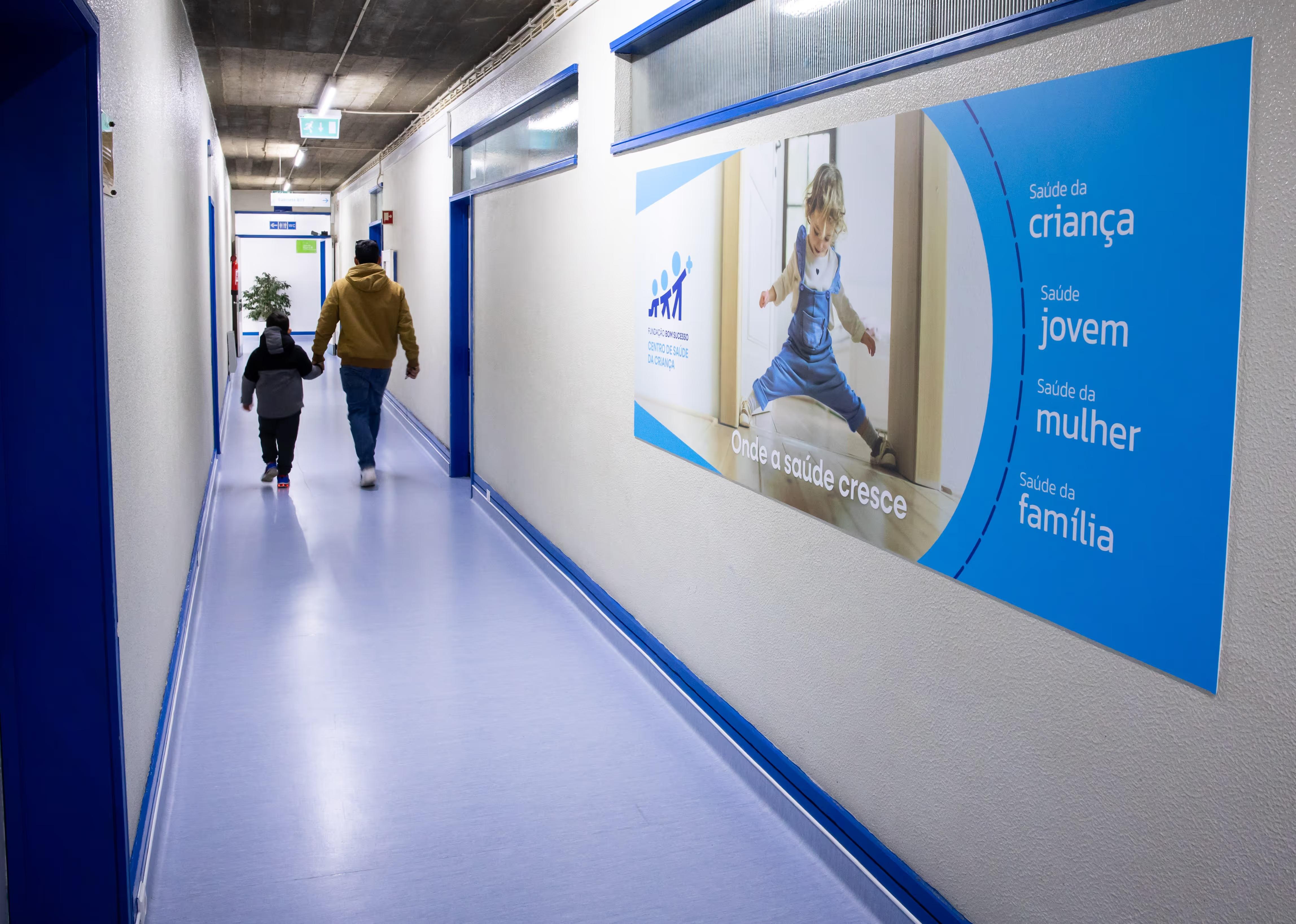 Man holding a child's hand walking down a hospital corridor with a health care poster on the wall.