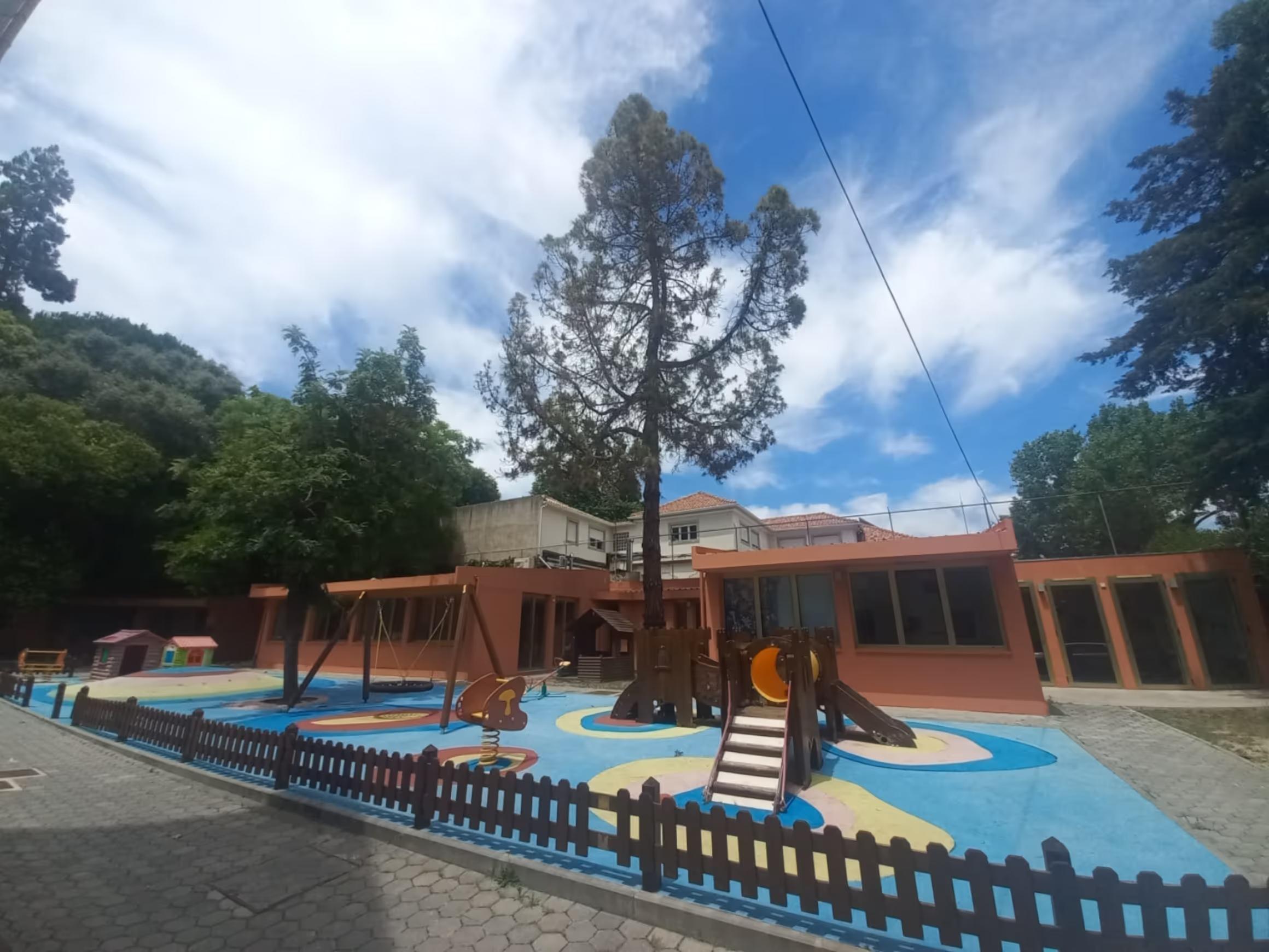 Outdoor playground with swings, slides, and playhouses surrounded by trees and a low fence.