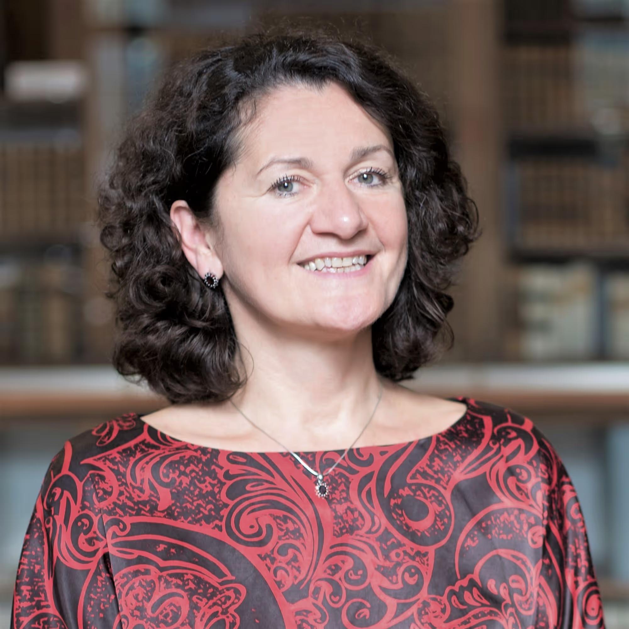 Smiling woman with curly dark hair wearing a red and black patterned top, standing indoors with blurred background.