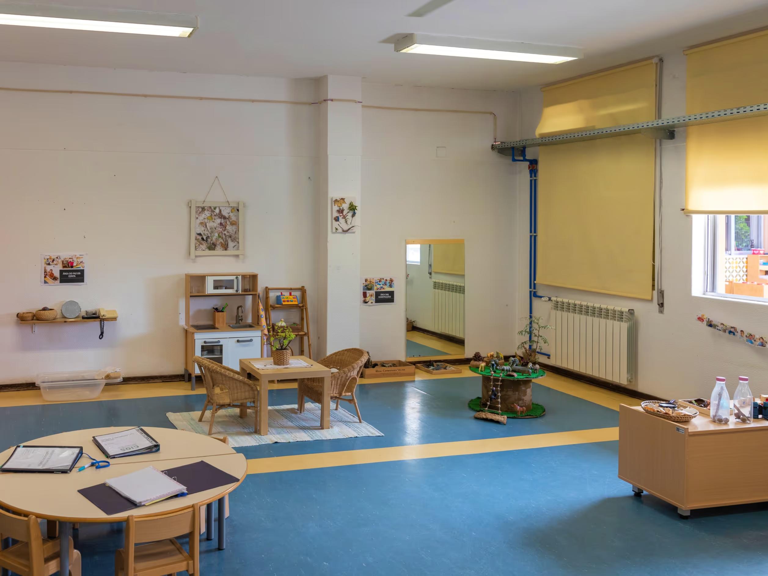 Bright classroom with small tables, chairs, play kitchen, mirror, and educational toys on blue and yellow floor.