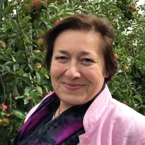 Smiling woman in a light pink jacket and dark scarf standing outdoors in front of leafy green trees with apples.