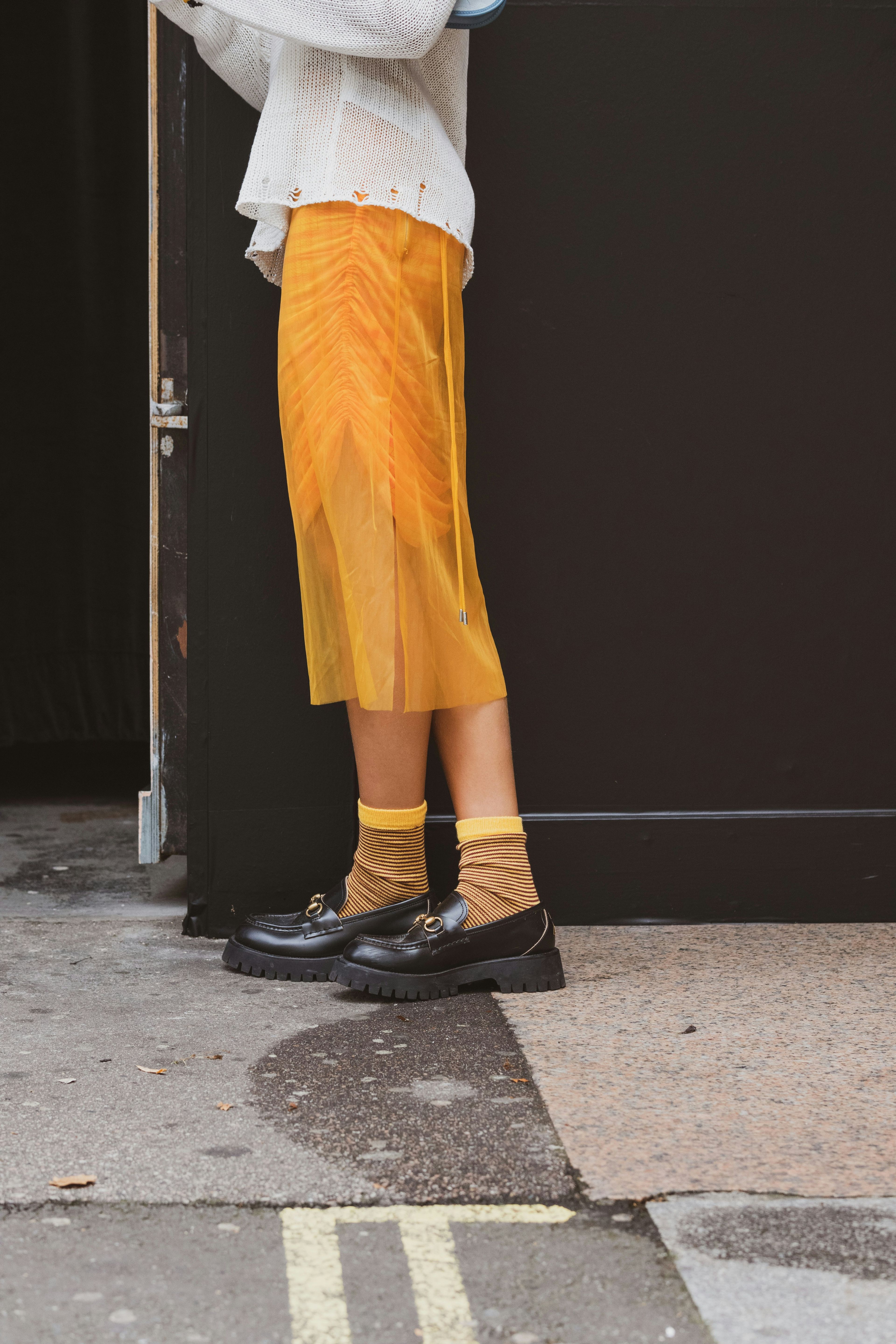 A close up of the lower half of someone's body, wearing black loafers and a yellow skirt with yellow socks, standing in front of a black wall with crossed legs.
