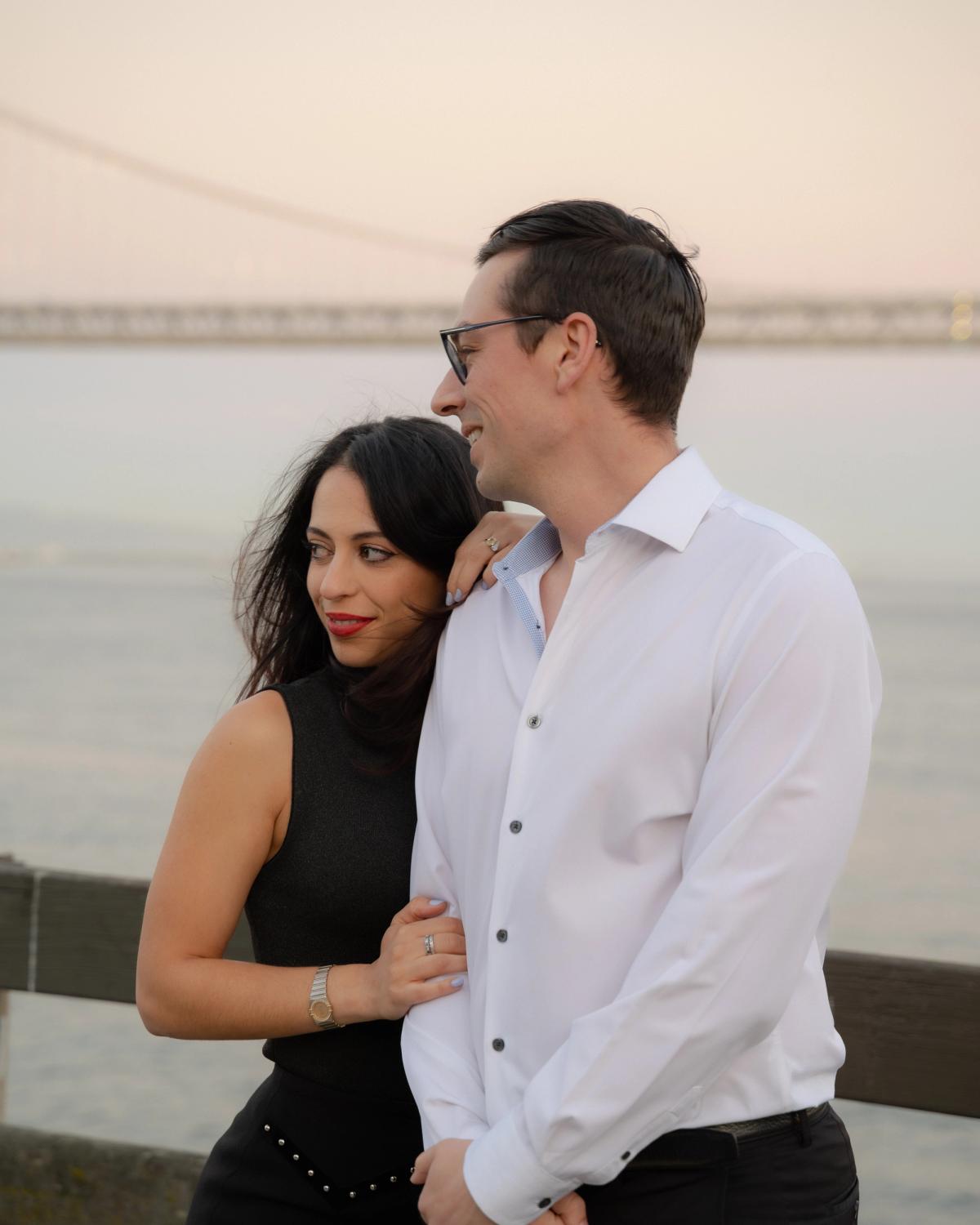 Engaged couple embracing on the waterfront at sunset with the Bay Bridge visible, San Francisco engagement photography