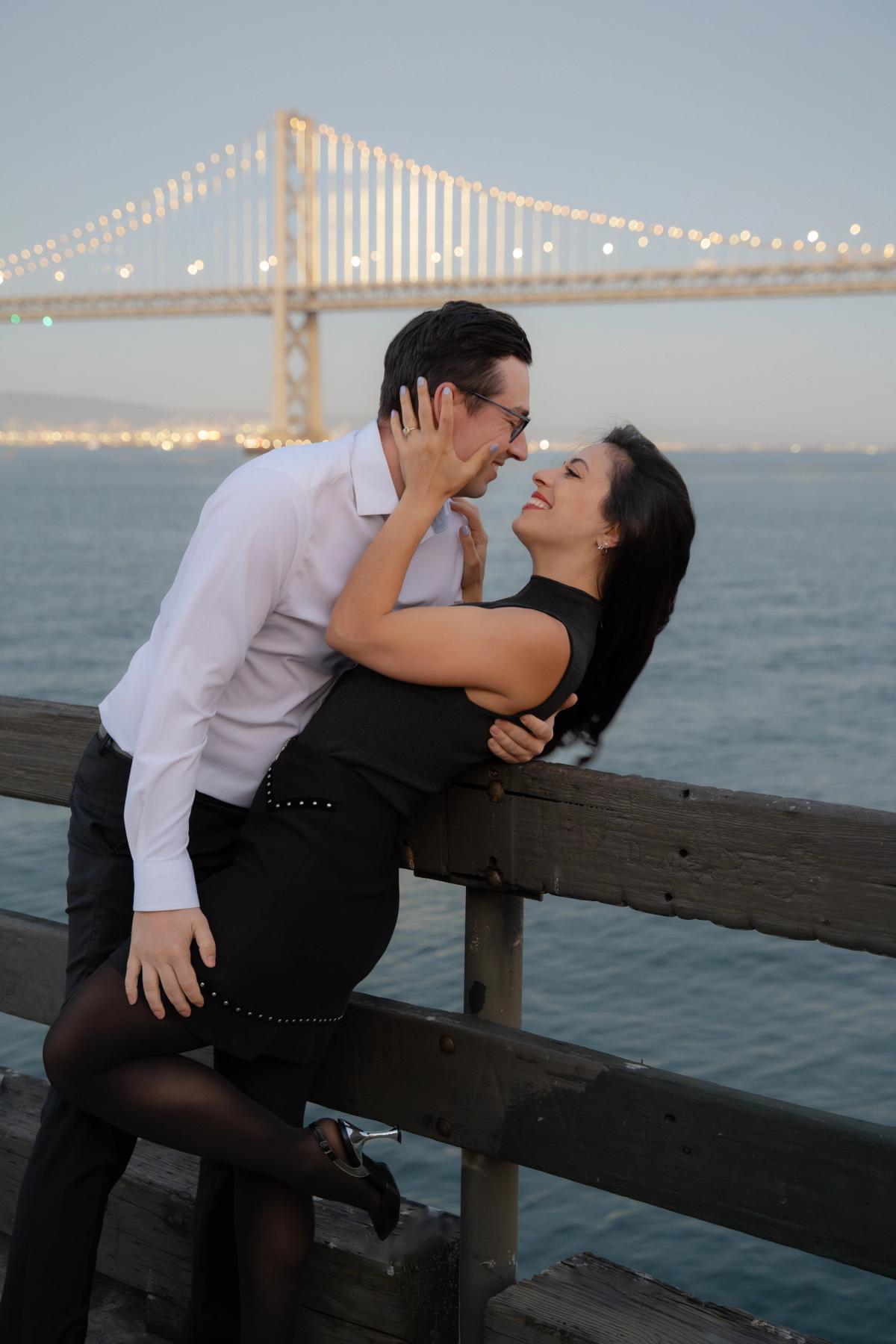 Couple leaning in for a romantic moment on a pier at twilight with the illuminated Bay Bridge glowing behind them, San Francisco engagement session