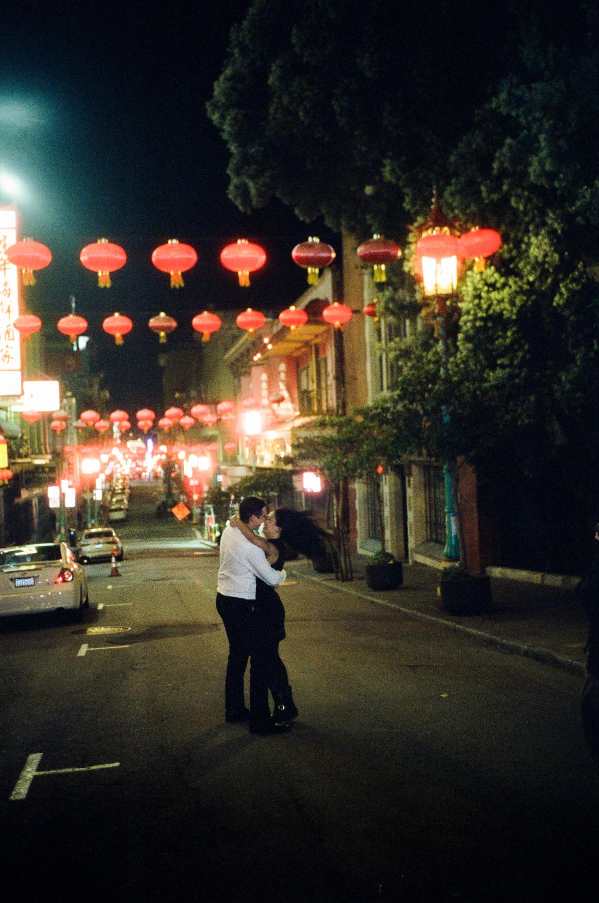 Couple embracing in the middle of a street at night beneath glowing red lanterns in San Francisco's Chinatown, film engagement photography