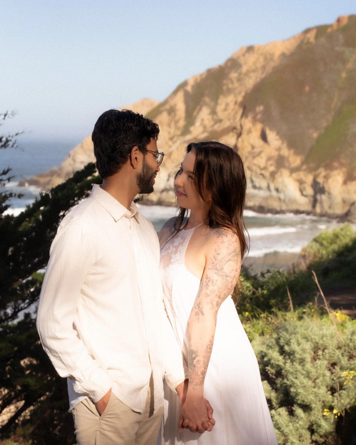 Couple holding hands and gazing at each other during Big Sur engagement session with rugged coastal cliffs and ocean behind them