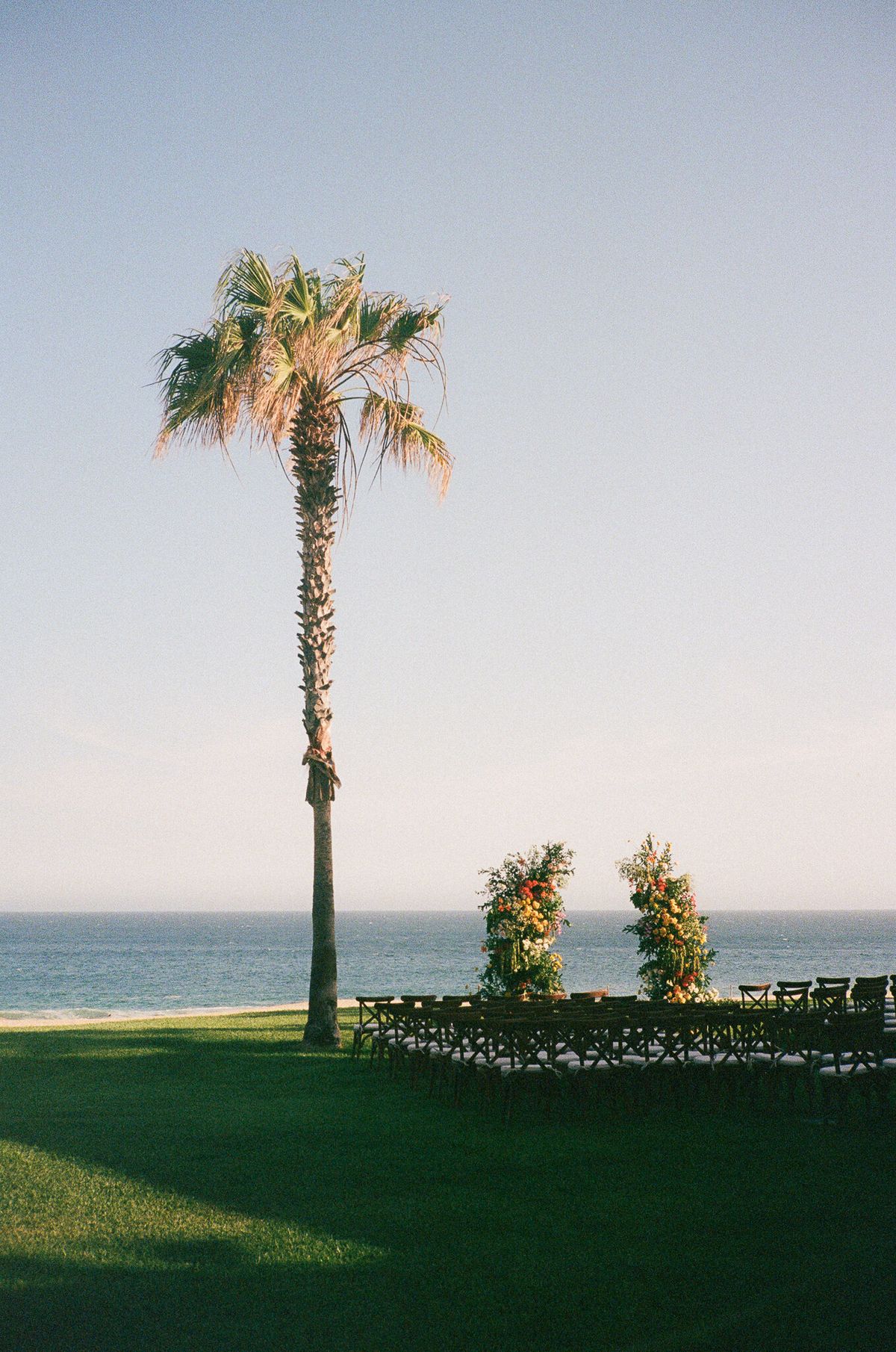 Oceanfront wedding ceremony setup at golden hour with floral pillars, cross-back chairs, and a palm tree silhouette against a clear sky