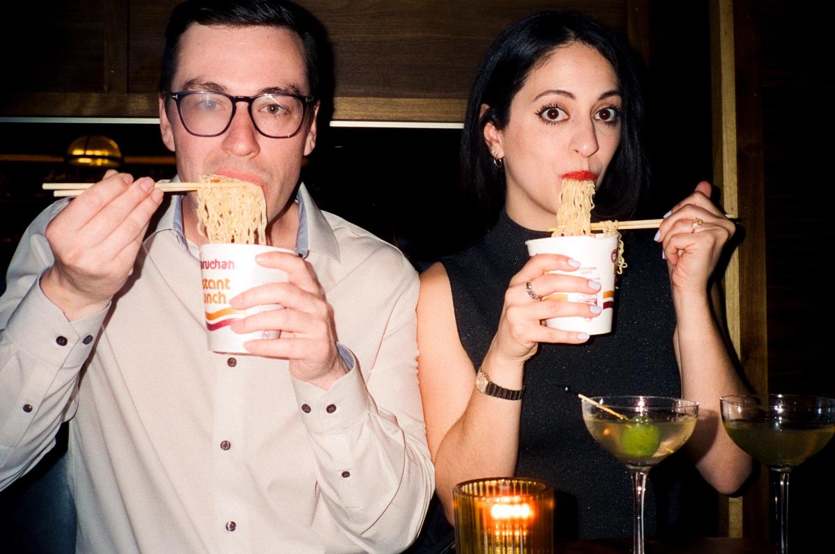 Couple eating instant ramen with chopsticks and making playful faces at a bar, fun candid engagement session moment