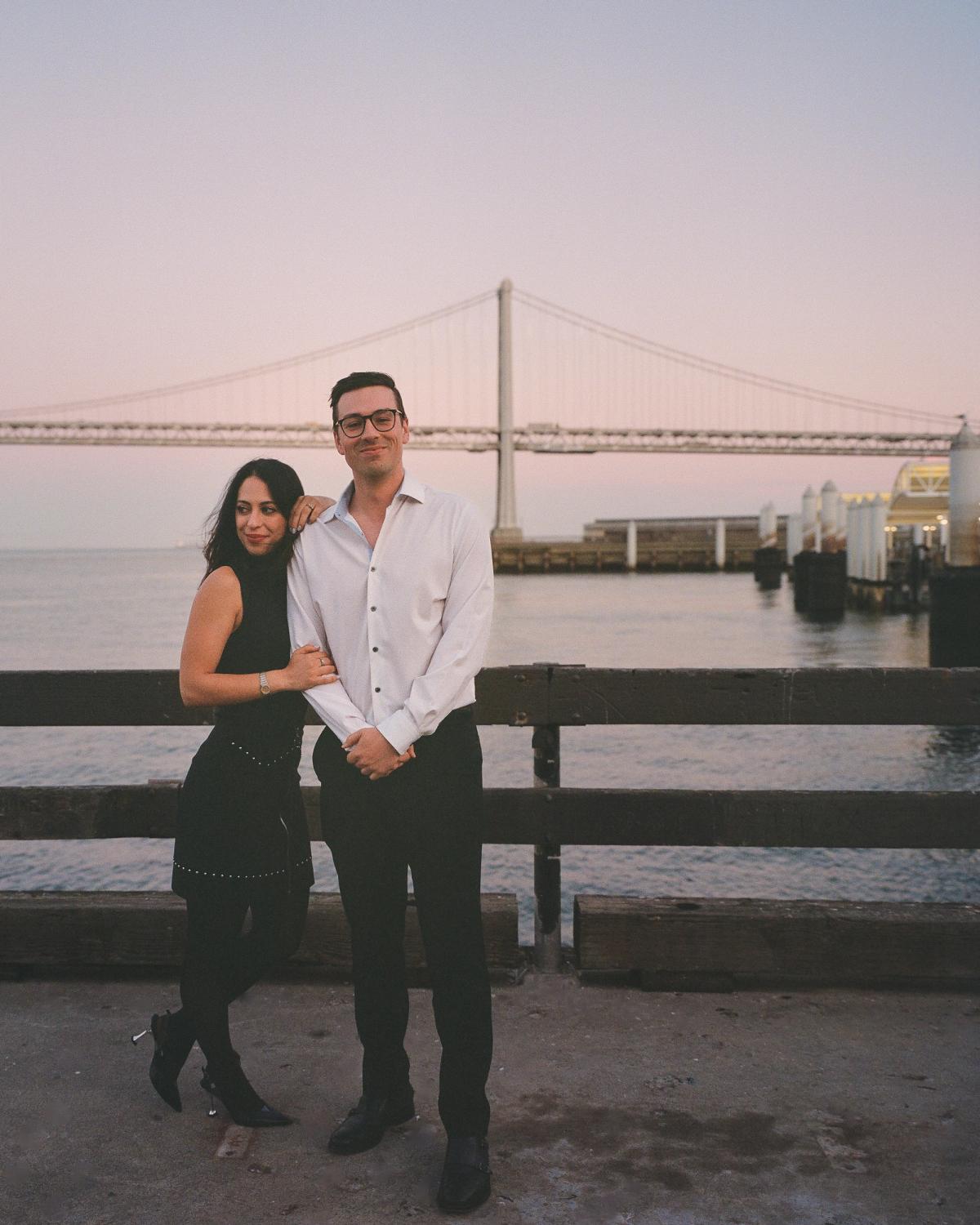 Couple posing together on the San Francisco waterfront at dusk with the Bay Bridge spanning the background, film photography engagement session