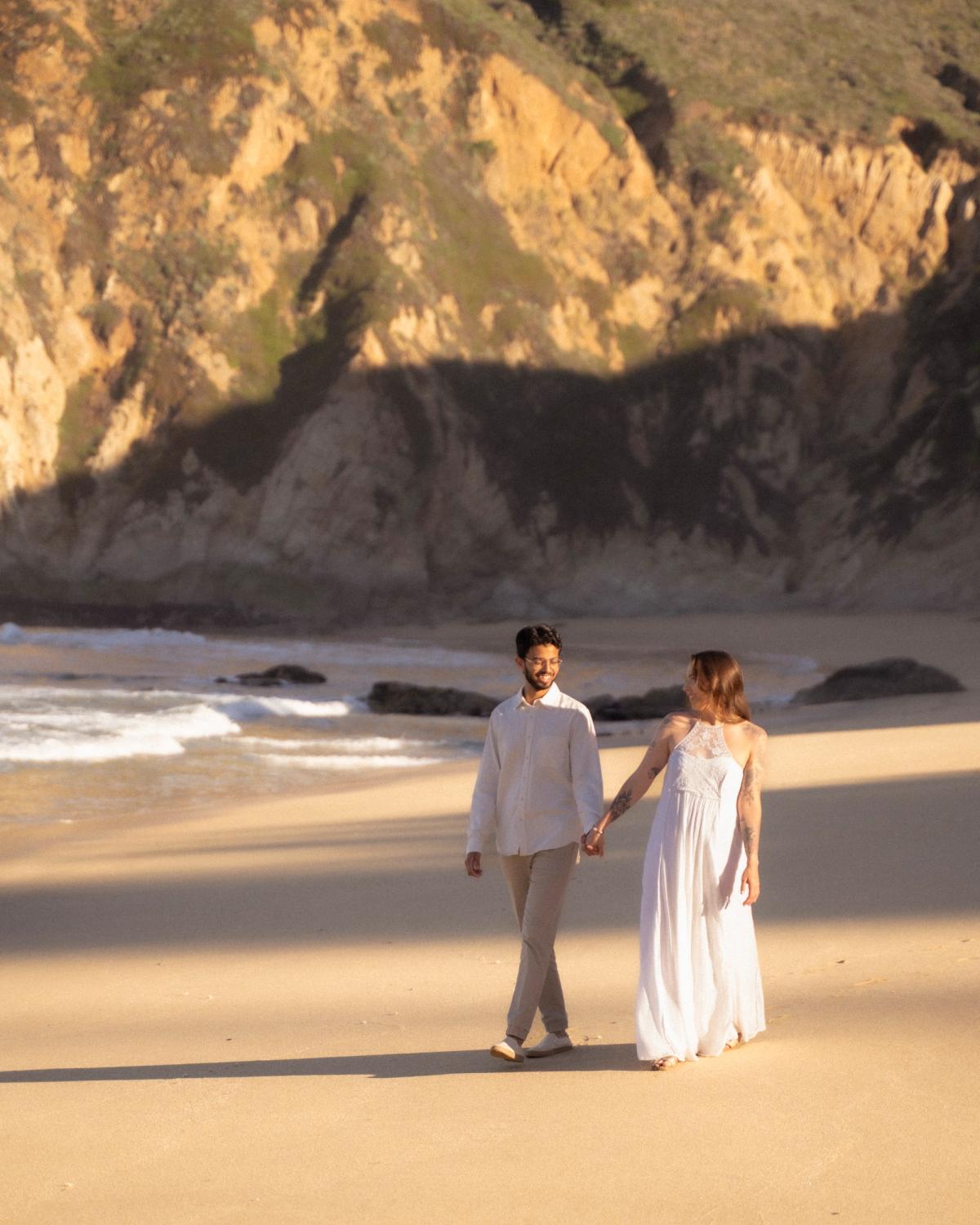 Engaged couple walking hand in hand on a sandy beach at golden hour with dramatic coastal cliffs in the background