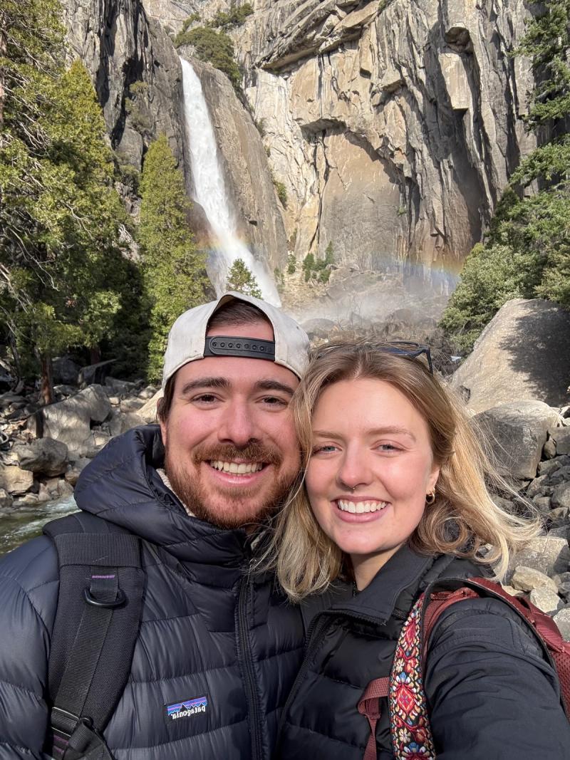Couple smiling together on a rocky overlook above a waterfall in Yosemite National Park, wearing hiking gear and backpacks