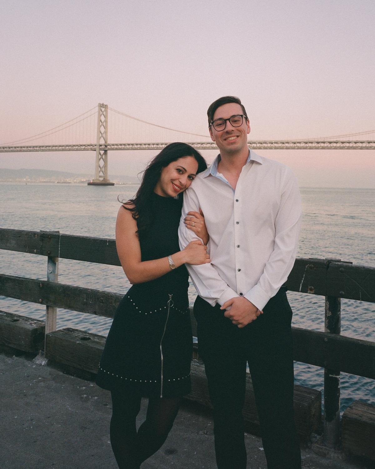 Smiling engaged couple standing together on the Embarcadero pier at sunset with the Bay Bridge behind them, film engagement photography