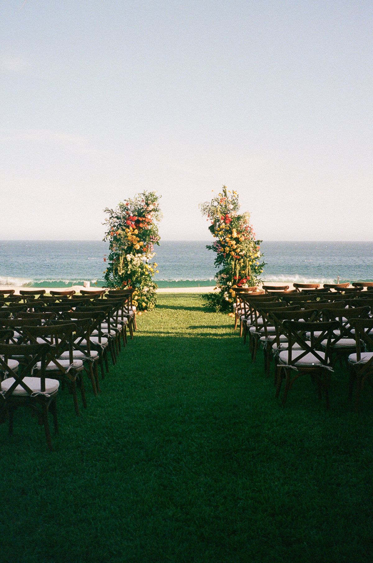 Outdoor beachfront wedding ceremony with tall floral arrangements and cross-back chairs, destination wedding setup