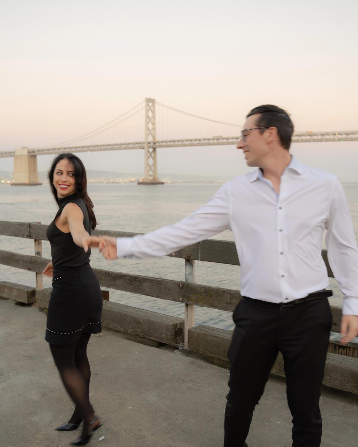 Couple dancing and laughing on the Embarcadero waterfront at dusk with the Bay Bridge in the background, San Francisco engagement session