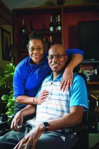 A black man in blue and white shirt sitting in a wheelchair with a black woman in blue shirt behind him.