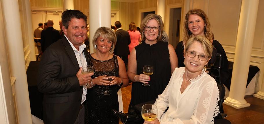 Four women and a man in formalwear hold wine glasses while posing for a photo at a fundraising event.