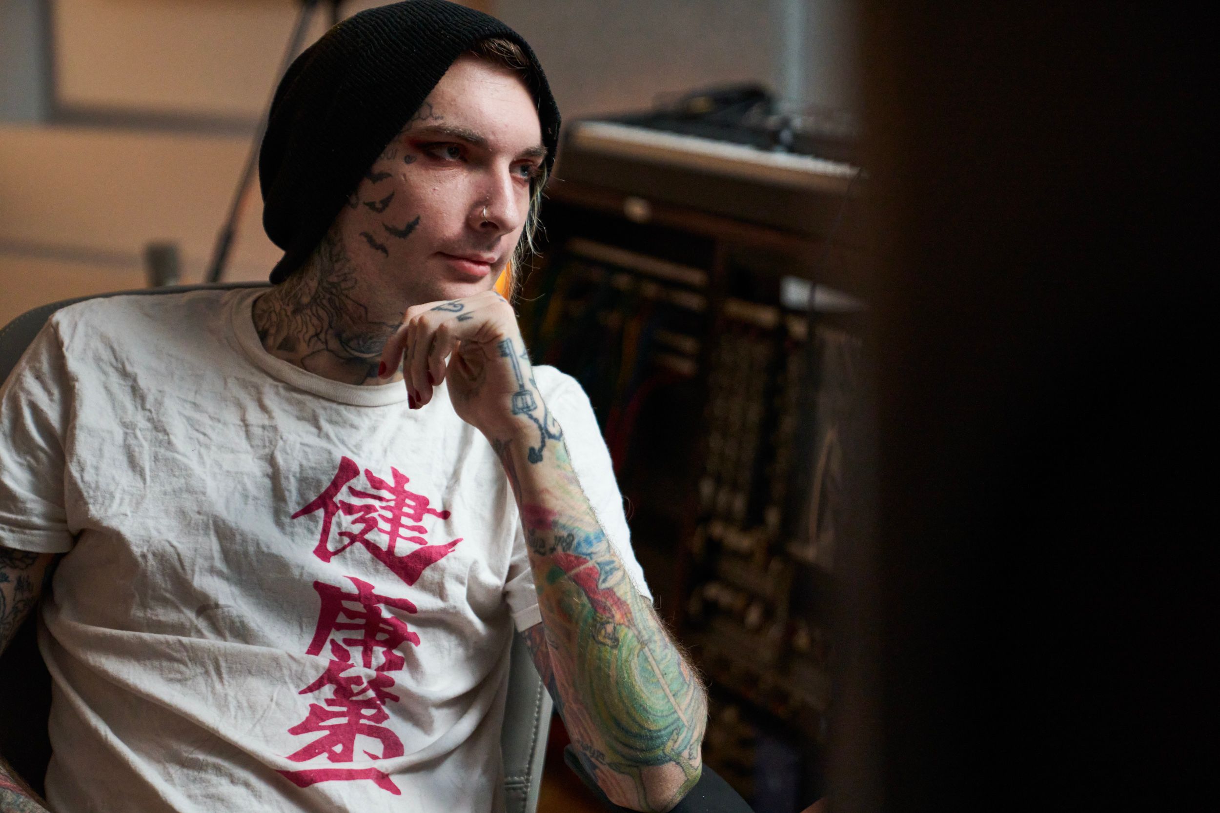 A young man with tattoos and a beanie rests his hand on his chin