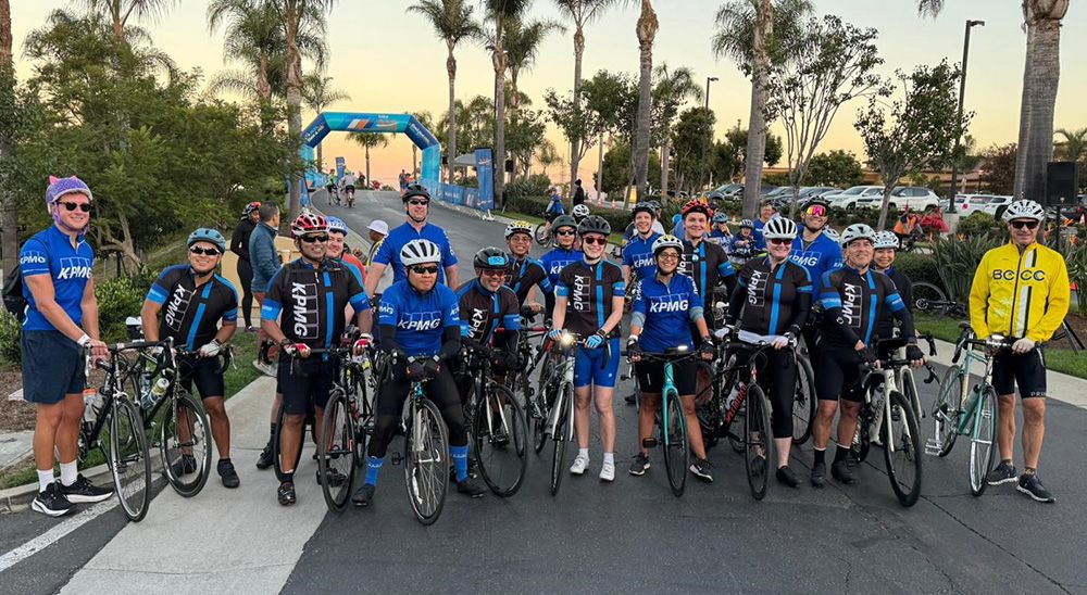 A group of cyclists in helmets and blue KPMG kits smile in front of palm trees and an inflated blue arch at the start of Bike MS.