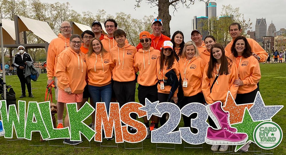 A group of people in orange shirts pose in front of large letters that say "Walk MS '23"