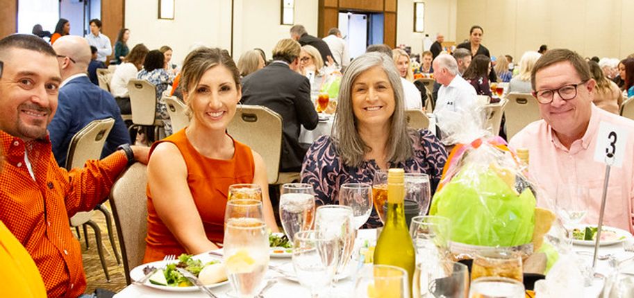 Two men and two women attending a fundraiser pose for a photo at a round table topped with dinner plates and wine glasses..