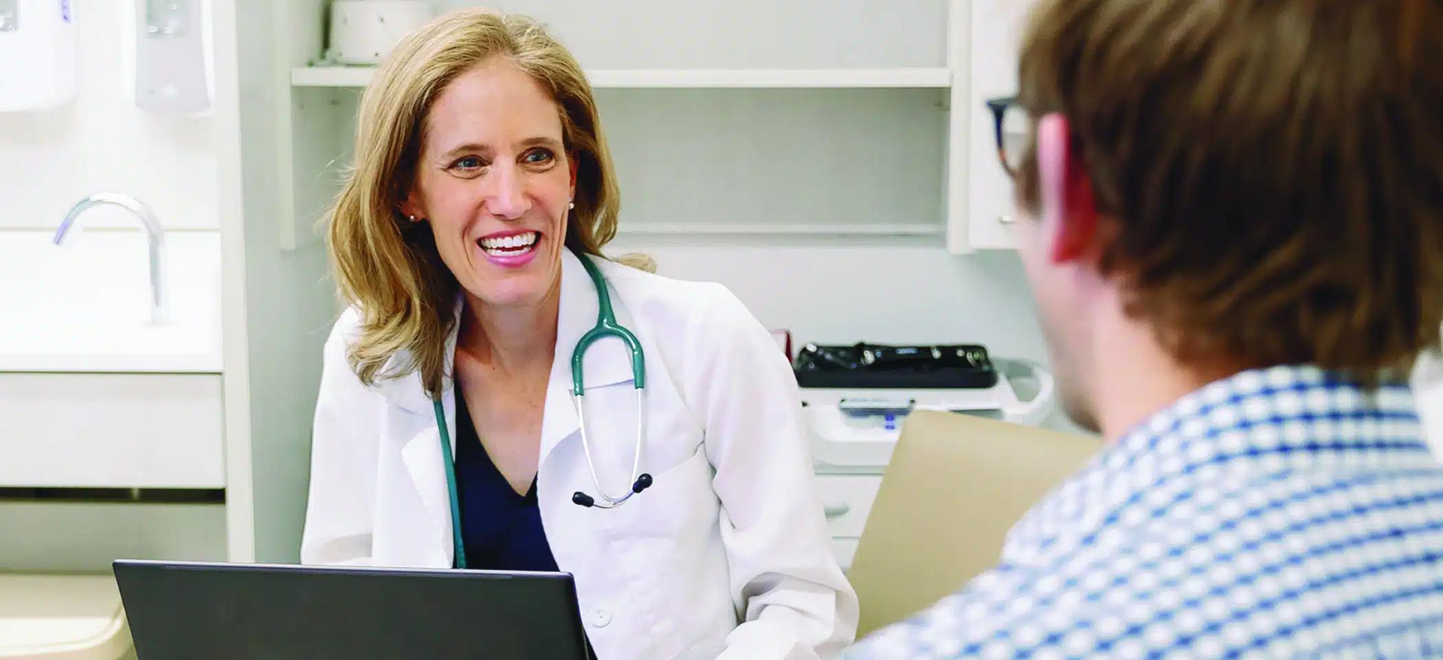 A blonde Caucasian woman in a white doctor's coat wears a stethoscope around her neck and has a laptop on her lap. Sitting in an exam room, she looks toward a patient, a man wearing glasses and a checkered shirt.