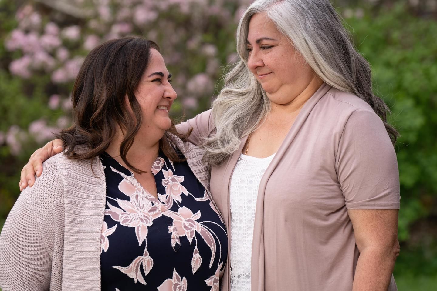 Heidi, a woman living with MS with long brown hair and a floral shirt, stands outside talking with and hugging a friend. 