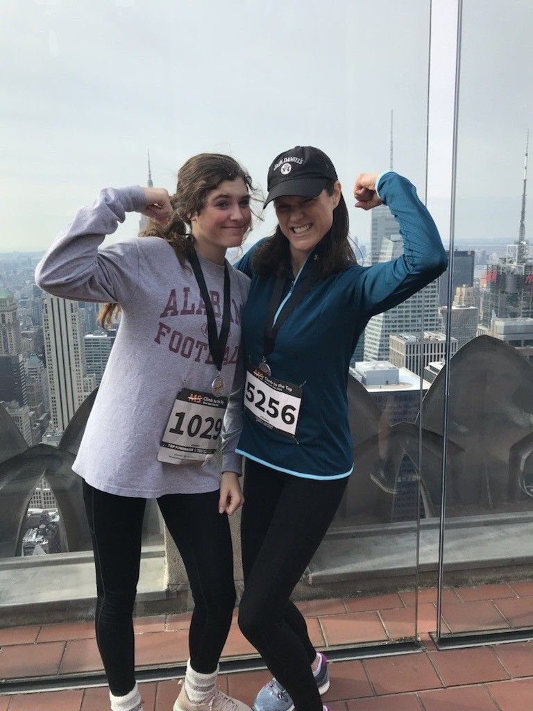Photo of a teenage daughter and her mother wearing racing bibs, pose in an embrace while showing their biceps at the top of Rockefeller Center, overlooking the Manhattan skyline. 
