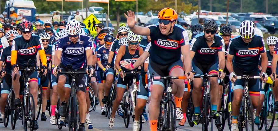 A group of cyclists in matching jerseys rides along the street, with banners indicating team names in the background.