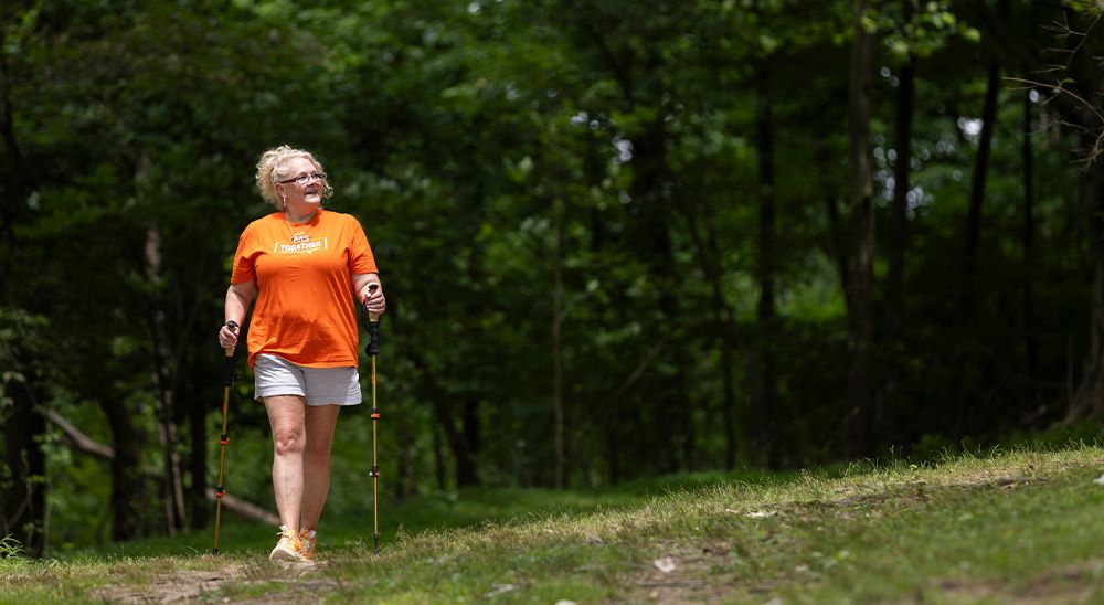 A woman wearing an orange shirt walks along a trail using walking sticks.