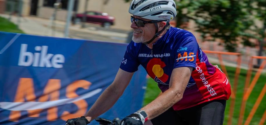 A cyclist wearing a blue, red and black cycling uniform rides past a Bike MS sign.