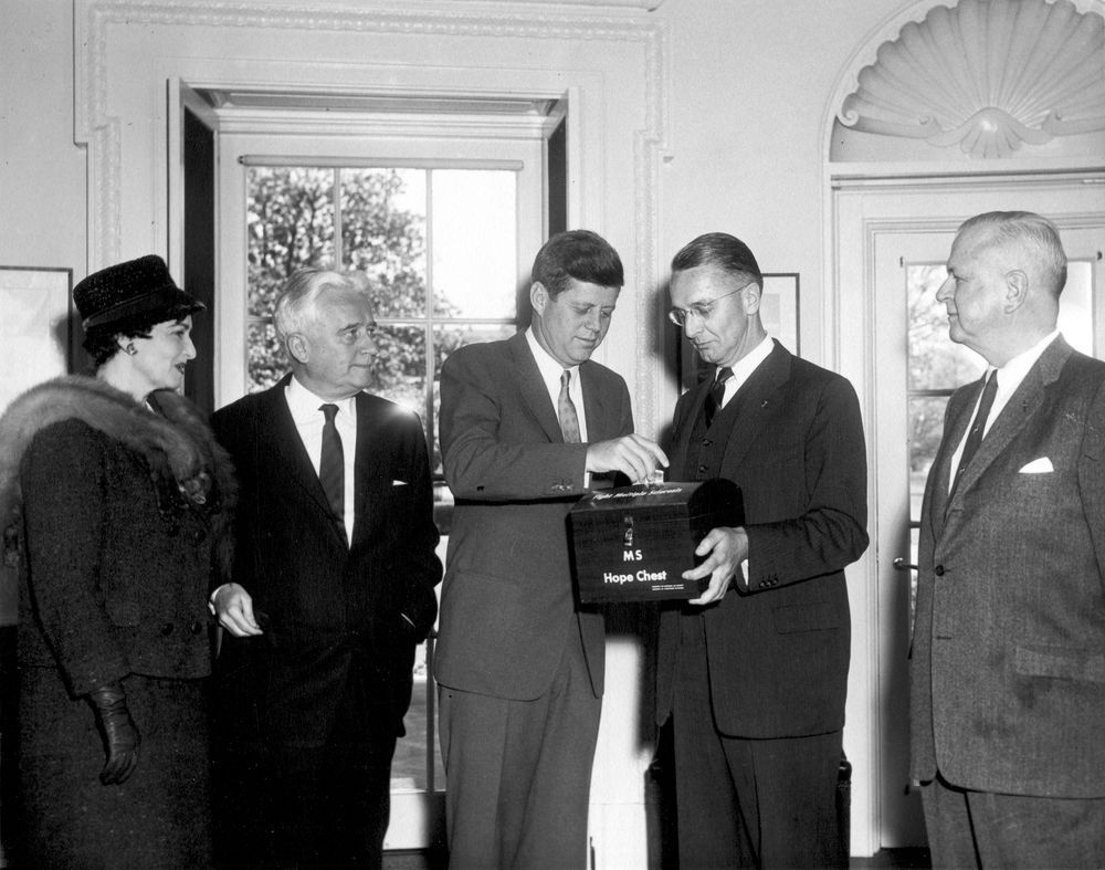 President John F. Kennedy adds money to a box labeled "MS Hope Chest." Sylvia Lawry and others stand beside him.