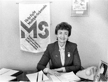 Sylvia Lawry sits at a desk. There is a banner hanging on the wall behind her her that says "Help fight Multiple Sclerosis."