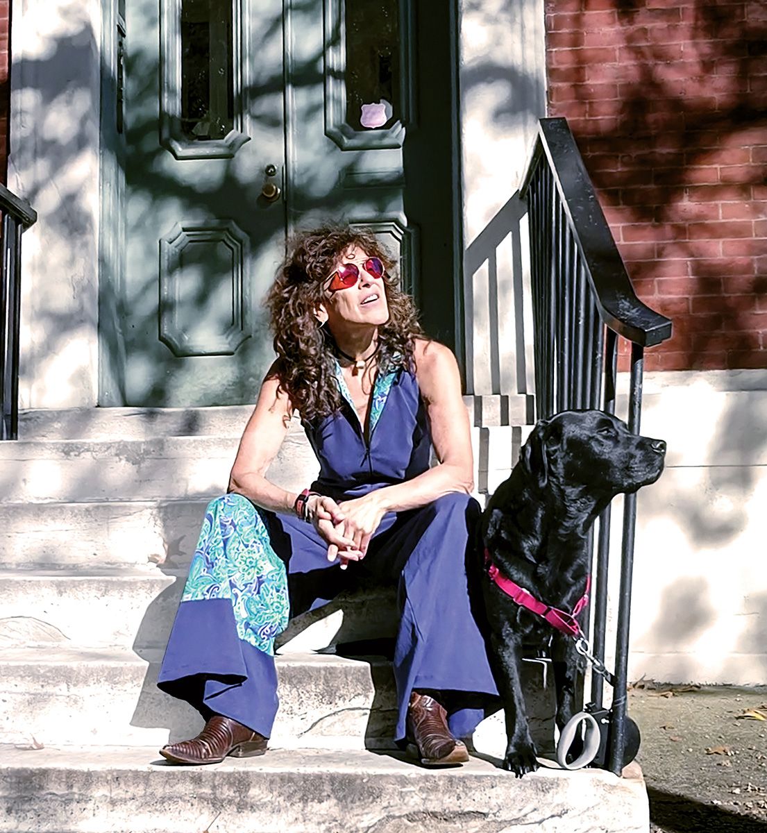 A woman wearing a jumpsuit sits on a stoop next to a dog.