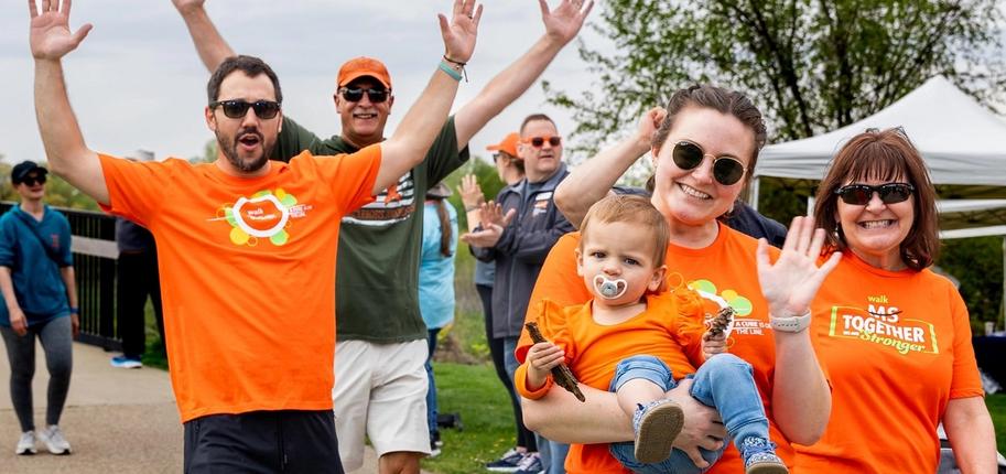Two women in orange shirts, one holding a baby in an orange shirt, and two men - one wearing orange, the other wearing green - smile and wave at a Walk MS event.