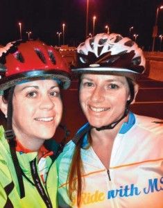 Two women in bike helmets and jerseys smile in a parking lot at night.