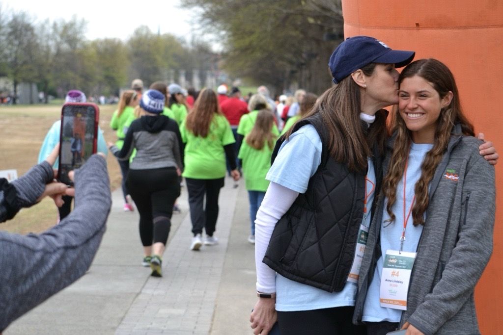 Photo of a mother embracing her smiling teenage daughter at a Walk MS event, with a group of people walking behind them.