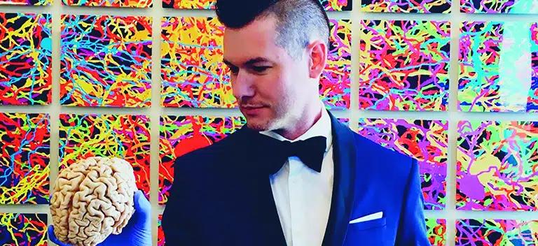 Shane Liddelow, PhD wearing a tuxedo and holding a human brain with a 3D image of brain pathways as the background