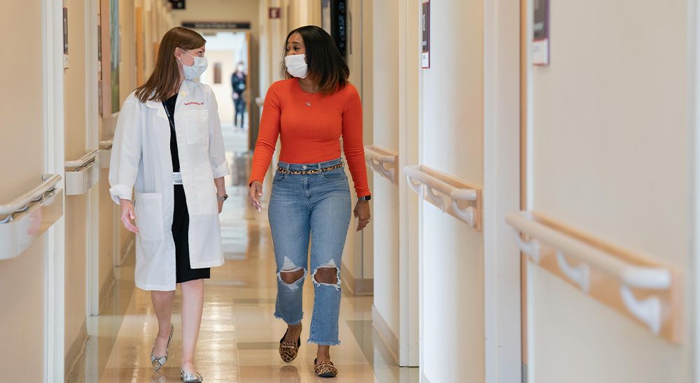 Two women walk down a hospital hallway in conversation. One is a doctor, wearing a white coat.