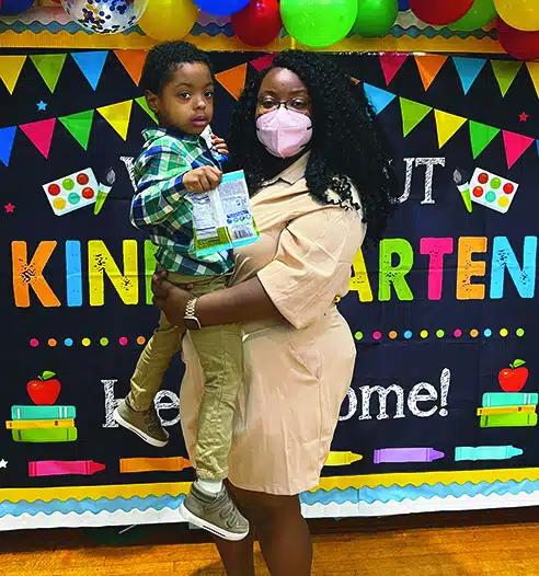 Moyna John-Caraballo, wearing a face mask and a tan dress, holds her son in her arms. They stand in front of a Kindergarten welcome sign.