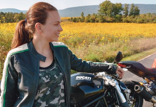 Diane Kramer stands outdoors, in front of a meadow and next to a Ducati motorcycle.