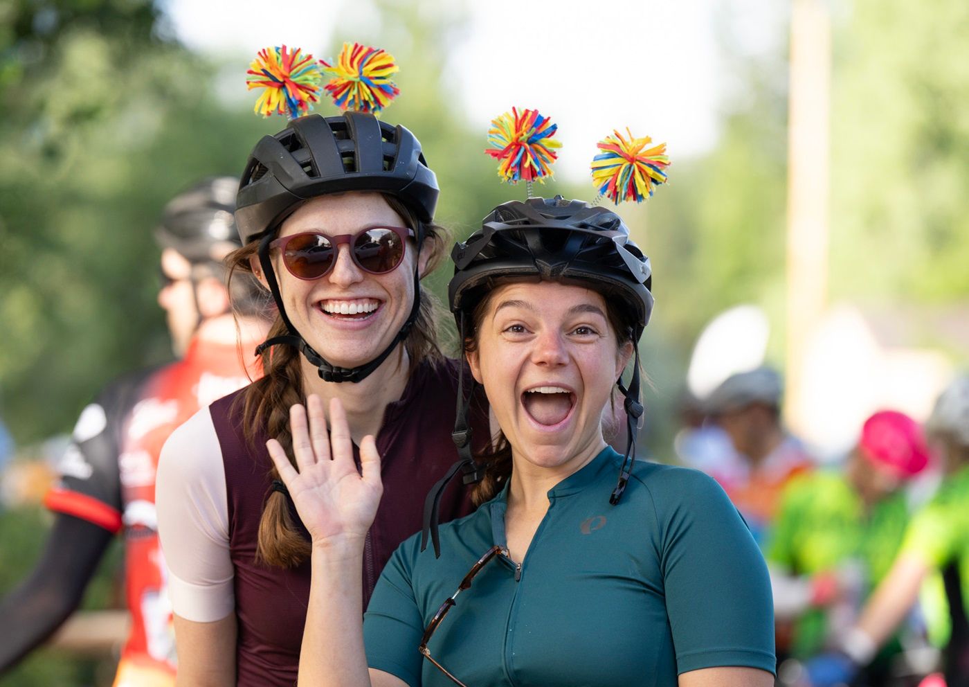 Two women wearing black bicycle helmets with colored pom-poms on top smile and wave.