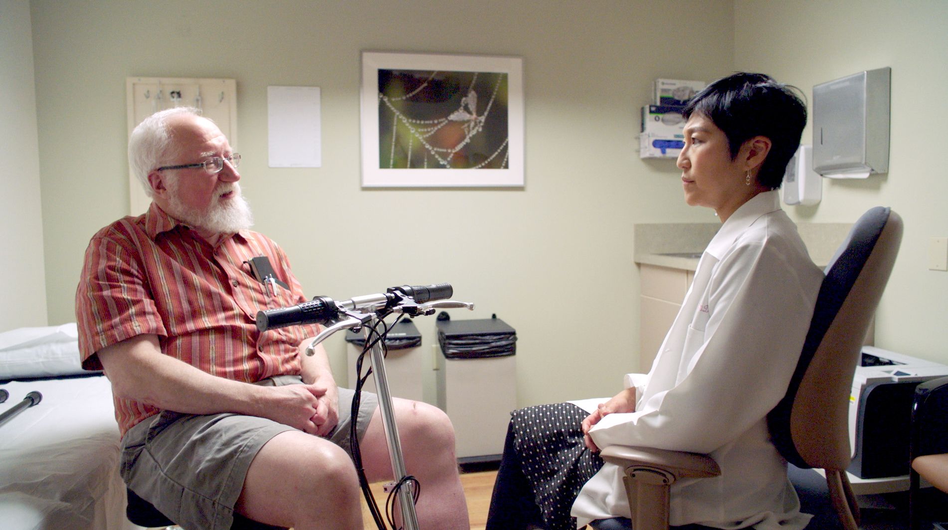 A man seated in a powerchair with white hair and a beard talks to a physician in a doctor’s office.