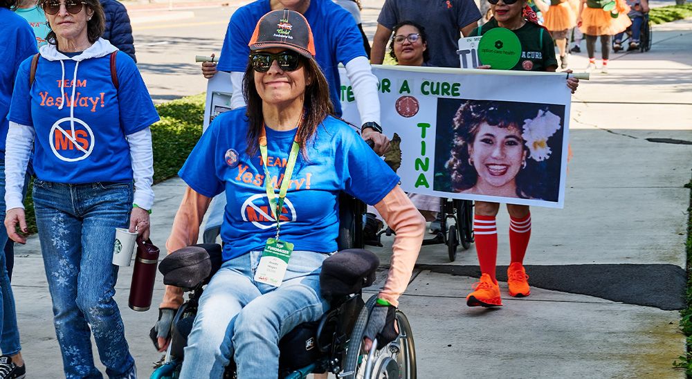A dark-haired woman in a wheelchair is pushed along a path by a fellow teammate.