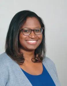 A black woman in glasses and wearing a blue shirt smiles at the camera.