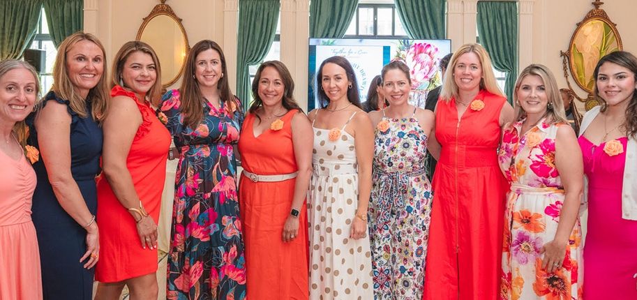 A group of 10 women stand side-by-side in orange, pink, white and blue dresses.