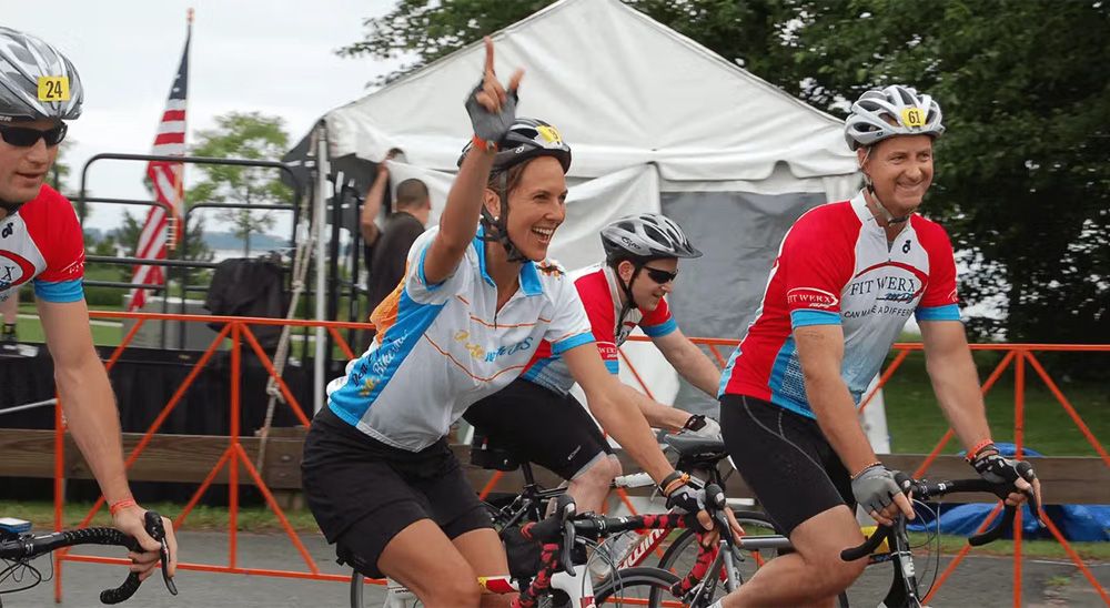 Four cyclists, including a woman in the middle who raises two fingers while smiling, ride together.