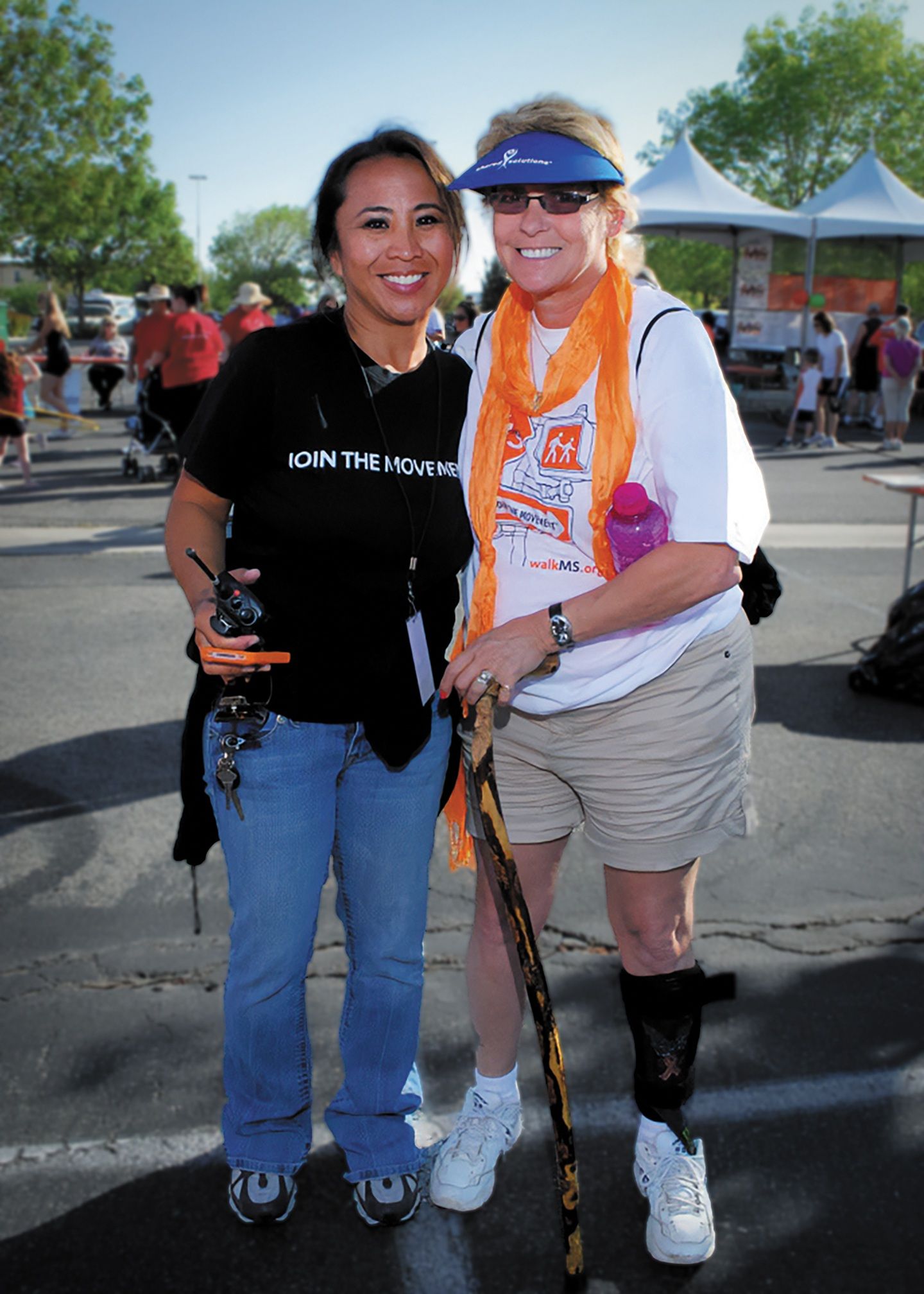 Woman in a black shirt and jeans, smiling softly while standing next to a woman in a white shirt and beige shorts.